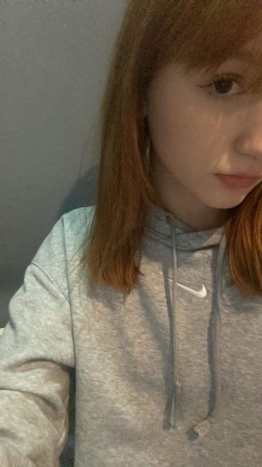 Portret młodej kobiety z rudymi włosami, ubrana w szarą bluzę z kapturem z logo Nike, z delikatnym makijażem oczu.