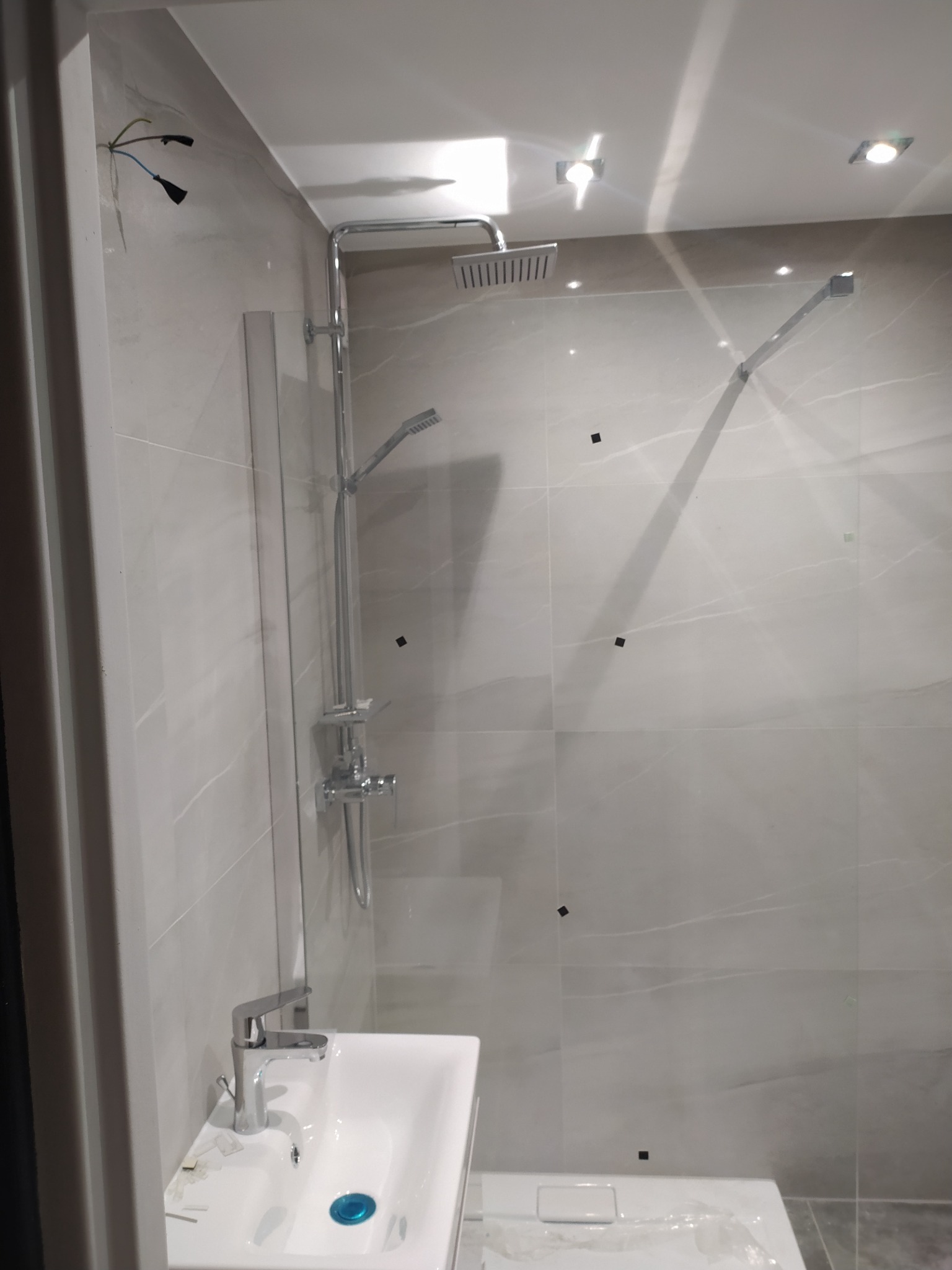 Nowoczesna łazienka z kabiną prysznicową typu walk-in, wykończona płytkami imitującymi marmur, z chromowaną armaturą i minimalistyczną umywalką, widoczne wystające kable elektryczne.