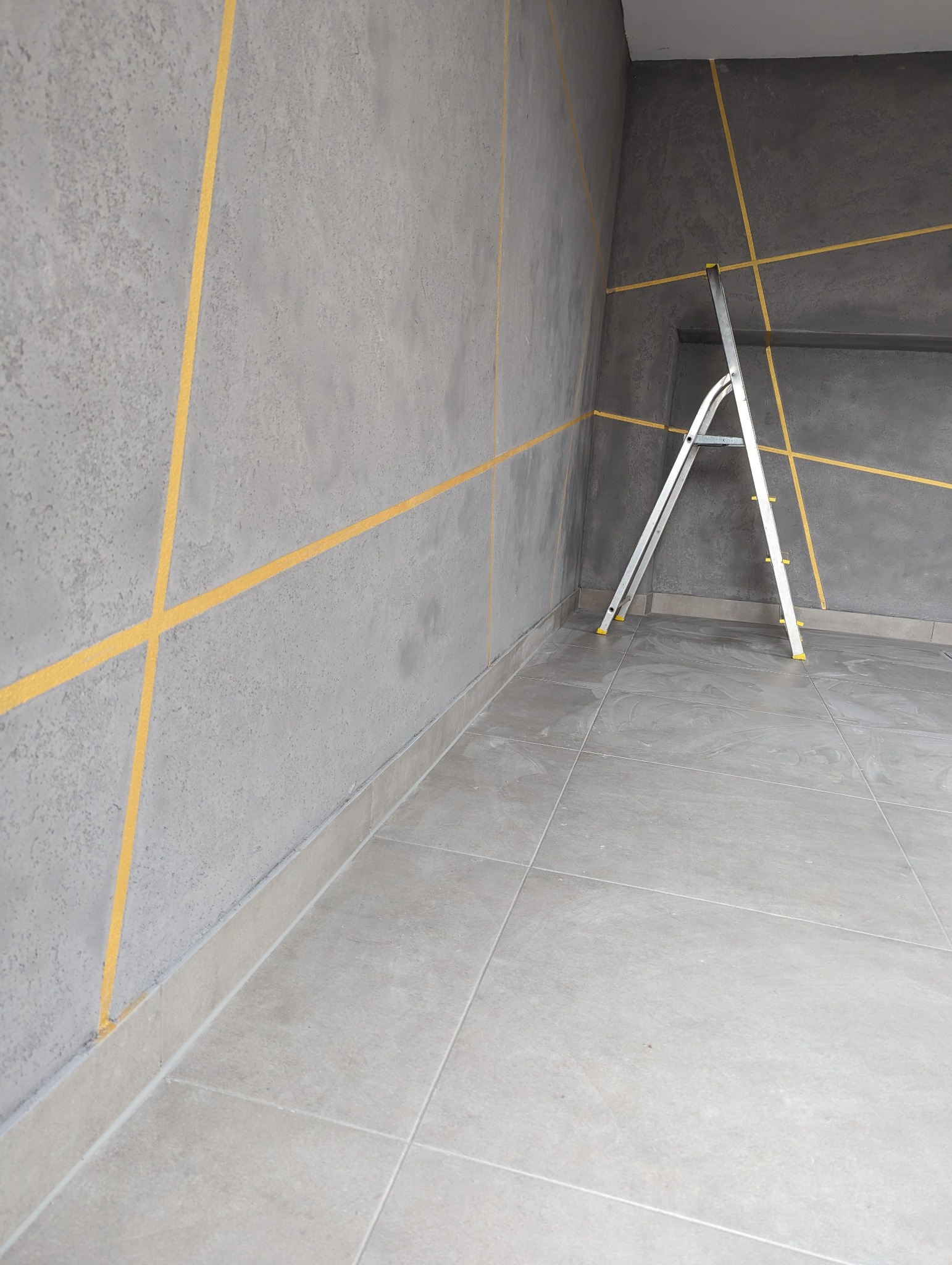 Szara ściana z geometrycznym wzorem z żółtej taśmy malarskiej, drabina aluminiowa oparta o ścianę, szara płytka podłogowa.