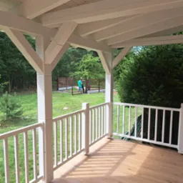 Zadaszony taras z drewnianą podłogą i białymi balustradami, widok na zielony ogród i osoby pracujące przy ogrodzeniu w tle.