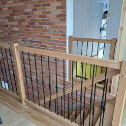 Ewa Krawczyk Zakład Stolarski KRAWMEBEL - Jasne, dębowe schody z czarnymi, metalowymi balustradami, widoczne z góry, z ceglaną ścianą w tle.