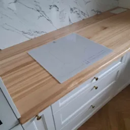 Jasny drewniany blat kuchenny z zamontowaną białą płytą indukcyjną Electrolux i białymi szafkami z trzema szufladami z ozdobnymi złotymi uchwytami, na tle marmurowej ściany.