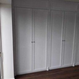 Ewa Krawczyk Zakład Stolarski KRAWMEBEL - Wbudowana szafa z białymi drzwiami panelowymi i srebrnymi gałkami, wykonana na wymiar w Ostródzie, widoczna na tle ciemnej drewnianej podłogi.