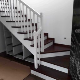 Ewa Krawczyk Zakład Stolarski KRAWMEBEL - Nowoczesne schody z ciemnego drewna z białymi balustradami i zabudową pod schodami, widoczne szuflady bez frontów.