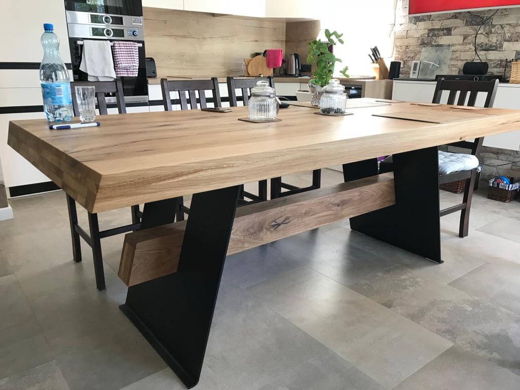 Duży, jasny stół z blatem z klejonego drewna o widocznej strukturze, osadzony na czarnych, metalowych nogach o trapezowym kształcie, z dodatkową półką z drewna pod blatem, w tle nowoczesna kuchnia.