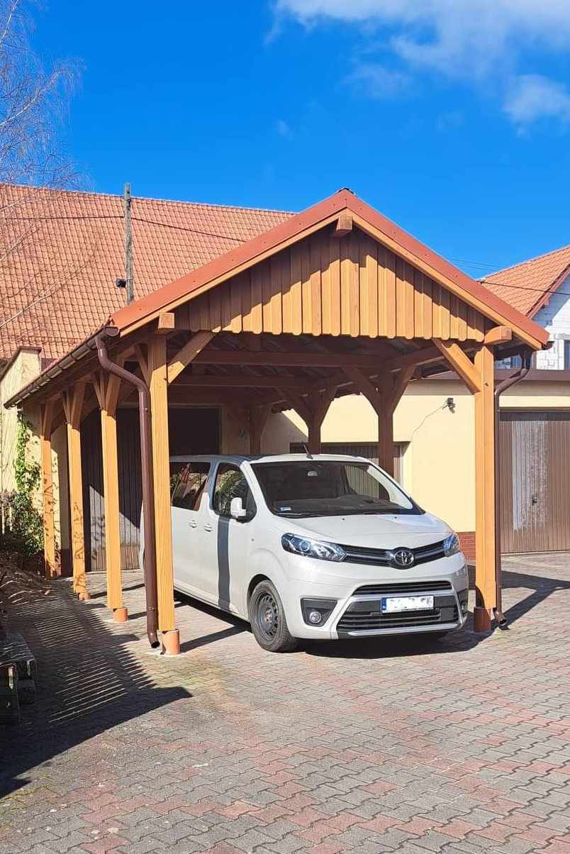 Drewniana wiata garażowa z brązowymi rynnami, chroniąca biały samochód na posesji z kostki brukowej w słoneczny dzień.