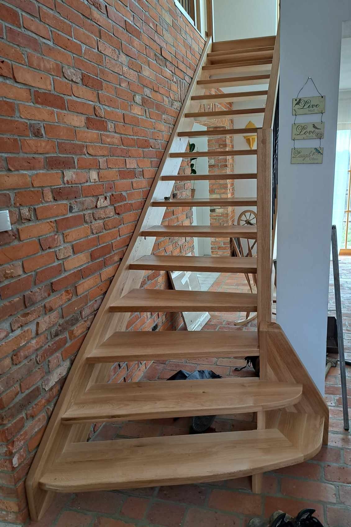 Nowoczesne, jasne schody z drewna dębowego, bez balustrady, przylegające do ściany z czerwonej cegły, z widoczną konstrukcją wspornikową stopni.
