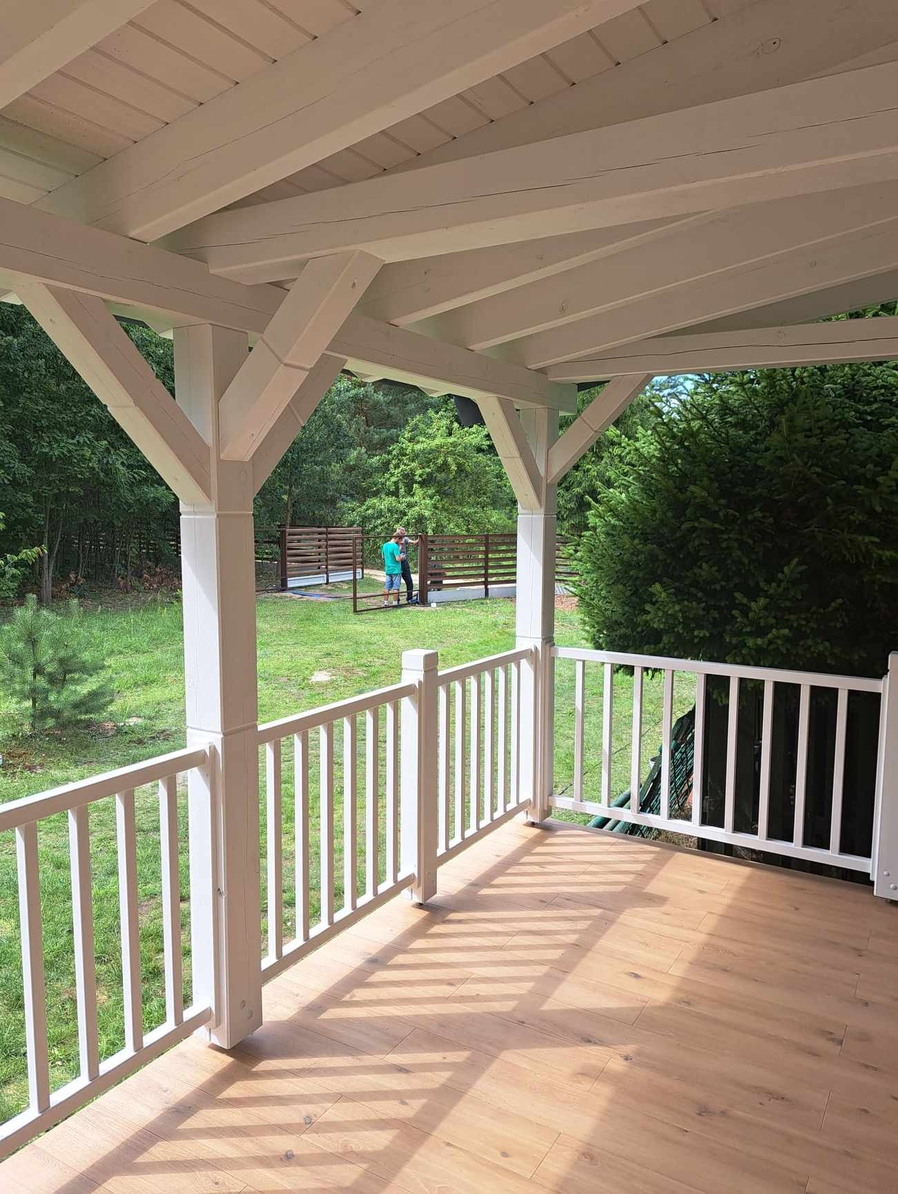 Zadaszony taras z drewnianą podłogą i białymi balustradami, widok na zielony ogród i osoby pracujące przy ogrodzeniu w tle.