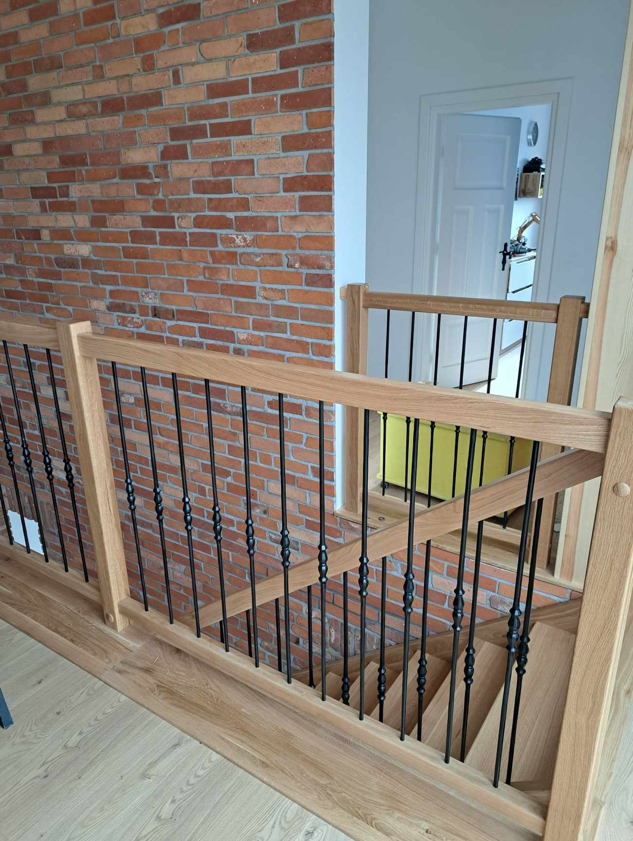 Jasne, dębowe schody z czarnymi, metalowymi balustradami, widoczne z góry, z ceglaną ścianą w tle.