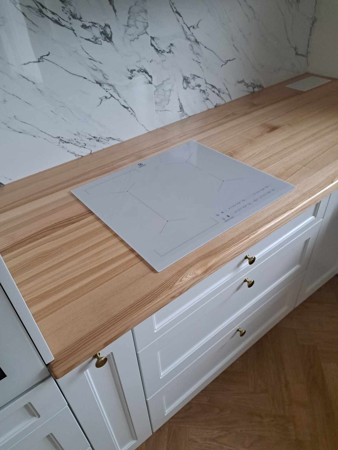 Jasny drewniany blat kuchenny z zamontowaną białą płytą indukcyjną Electrolux i białymi szafkami z trzema szufladami z ozdobnymi złotymi uchwytami, na tle marmurowej ściany.