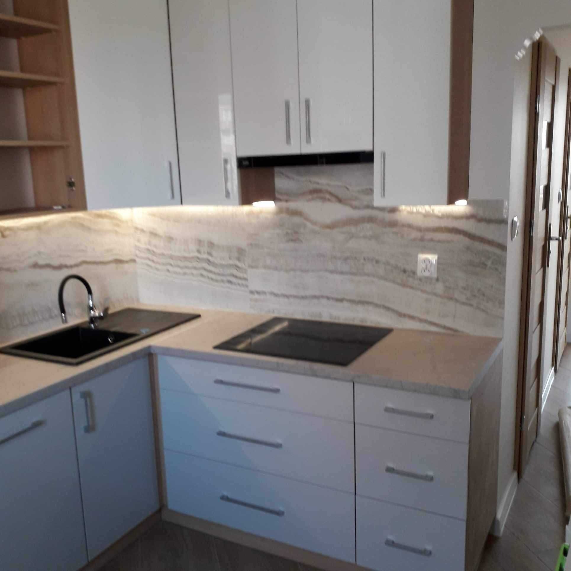 Nowoczesna kuchnia z białymi frontami szafek, czarnym zlewem i płytą indukcyjną, oraz blatem imitującym marmur. Podświetlenie LED podkreśla unikalny wzór na ścianie nad blatem.