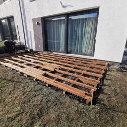 AP Invest - Montaż drewnianej konstrukcji tarasu na czarnej membranie przy nowoczesnym domu z dużymi oknami; widoczny pracownik w tle, wczesna faza budowy.