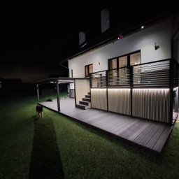 AP Invest - Nowoczesny taras kompozytowy z oświetleniem LED i pergolą, widoczny w nocy z psem na trawniku.