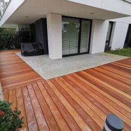 AP Invest - Nowoczesny taras z desek w odcieniach brązu, przylegający do budynku z dużymi oknami, oświetlony lampą ogrodową i dekorowany zimozielonym krzewem z czerwonymi owocami.