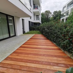 AP Invest - Nowoczesny taras z desek w odcieniach brązu, przylegający do budynku apartamentowca, z widoczną roślinnością oddzielającą go od sąsiedniego budynku.