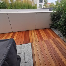 AP Invest - Ułożony taras z desek w odcieniach brązu, częściowo przykryty pokrowcem, z widokiem na zieleń i fragment budynku w tle.