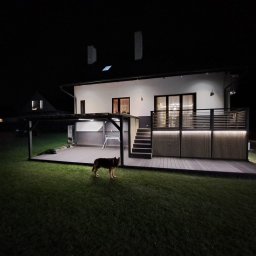AP Invest - Oświetlony taras z kompozytu przy nowoczesnym domu nocą, z psem na trawniku i zadaszeniem z boku.