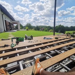 AP Invest - Montaż desek tarasowych na aluminiowej konstrukcji, widok z perspektywy osoby siedzącej na tarasie, narzędzia i akcesoria budowlane w tle, nowoczesny dom z zielonym trawnikiem i drzewami w tle.