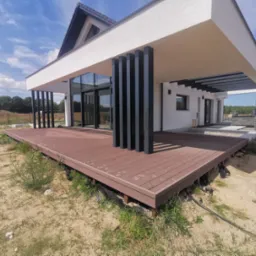 Nowoczesny dom z przestronnym tarasem z desek kompozytowych w kolorze brązu, z czarnymi filarami podtrzymującymi zadaszenie, widok na ogród i błękitne niebo.