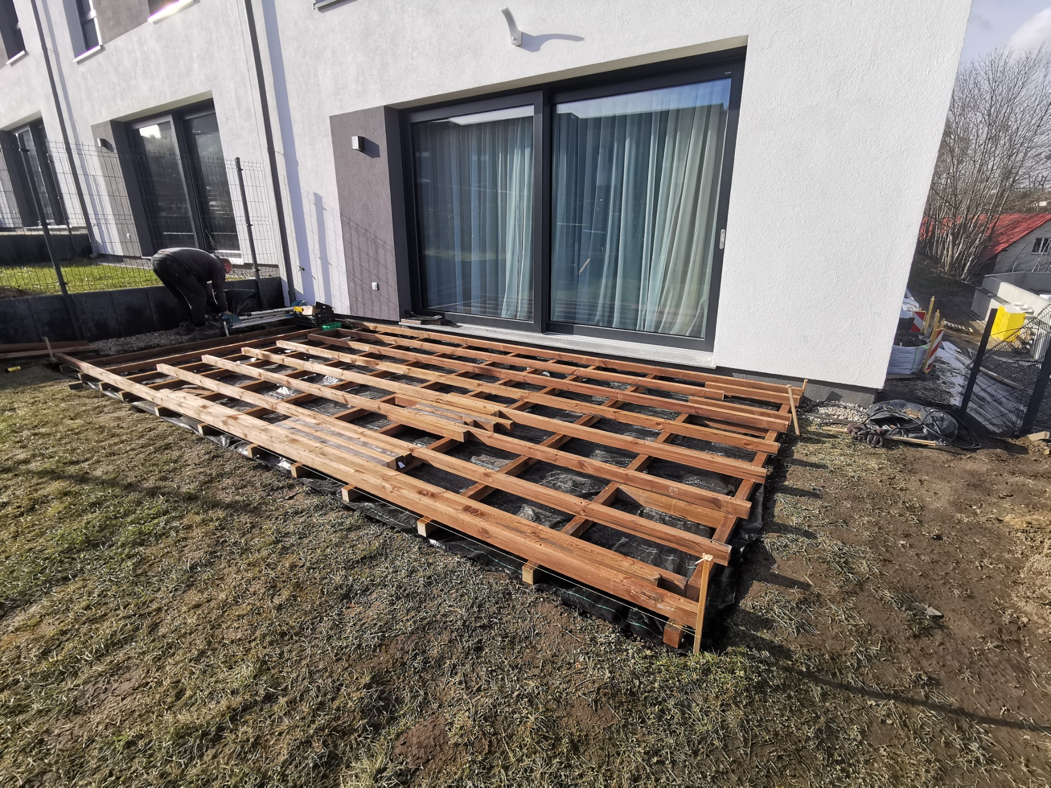 Montaż drewnianej konstrukcji tarasu na czarnej membranie przy nowoczesnym domu z dużymi oknami; widoczny pracownik w tle, wczesna faza budowy.