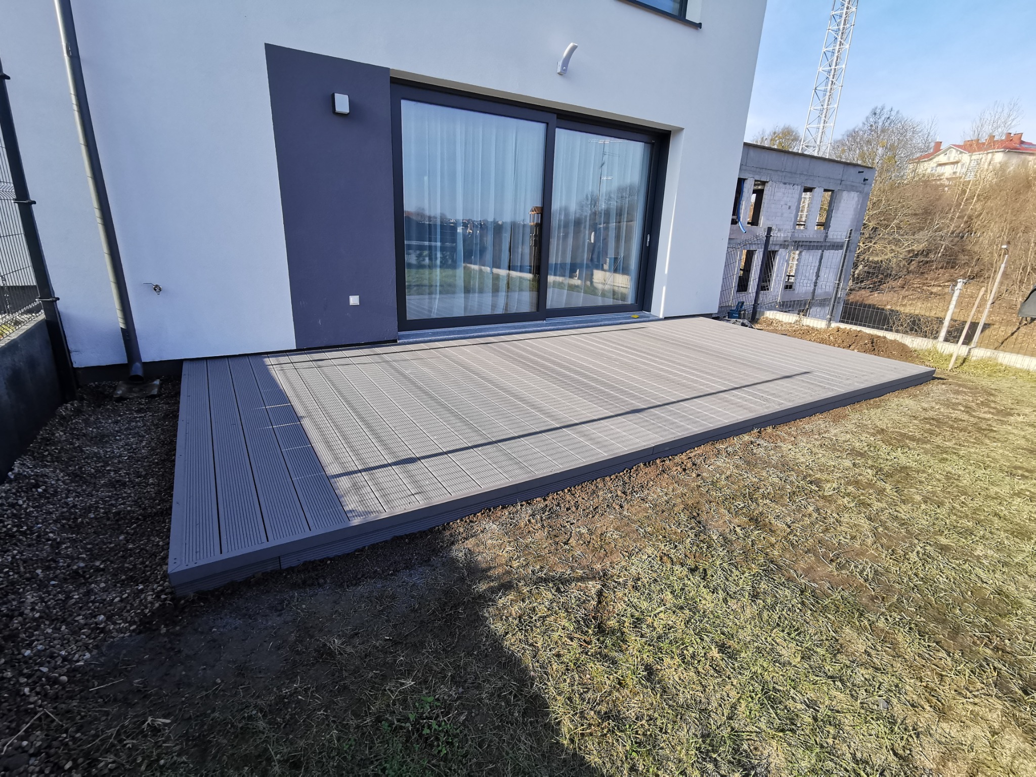 Szary taras z desek kompozytowych przylegający do nowoczesnego domu z dużymi przesuwnymi drzwiami tarasowymi, widok na plac budowy w tle.