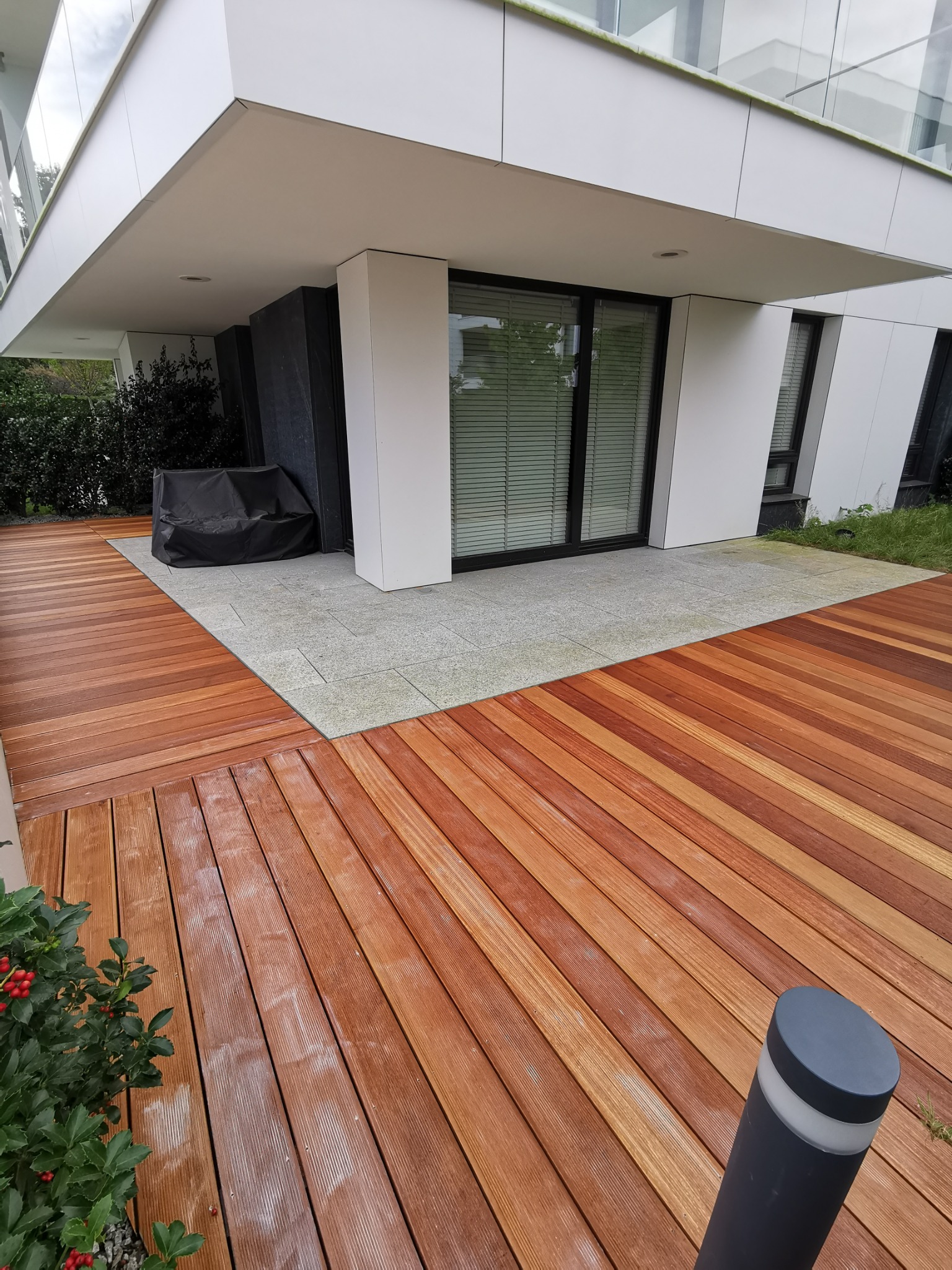 Nowoczesny taras z desek w odcieniach brązu, przylegający do budynku z dużymi oknami, oświetlony lampą ogrodową i dekorowany zimozielonym krzewem z czerwonymi owocami.