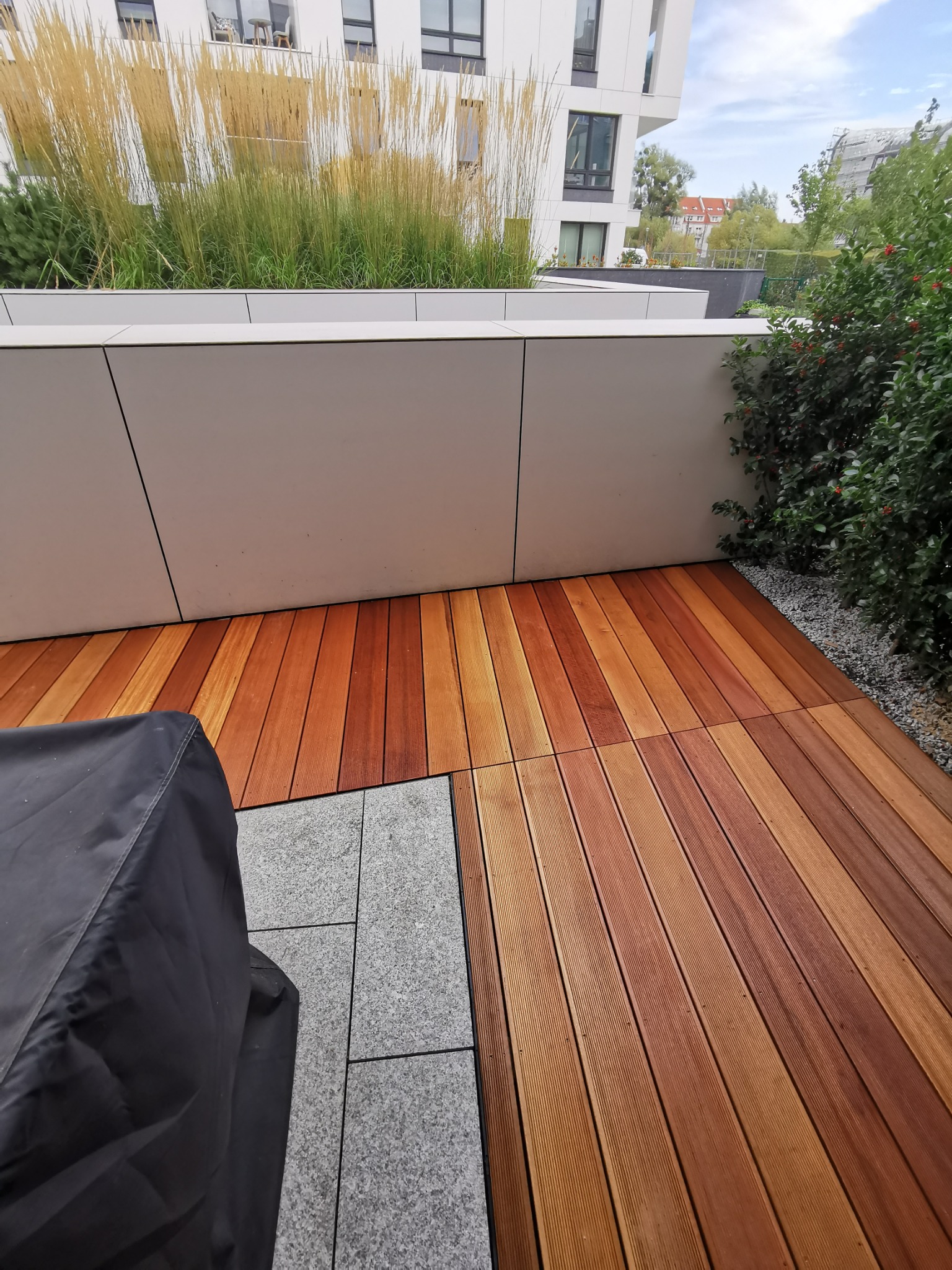 Ułożony taras z desek w odcieniach brązu, częściowo przykryty pokrowcem, z widokiem na zieleń i fragment budynku w tle.