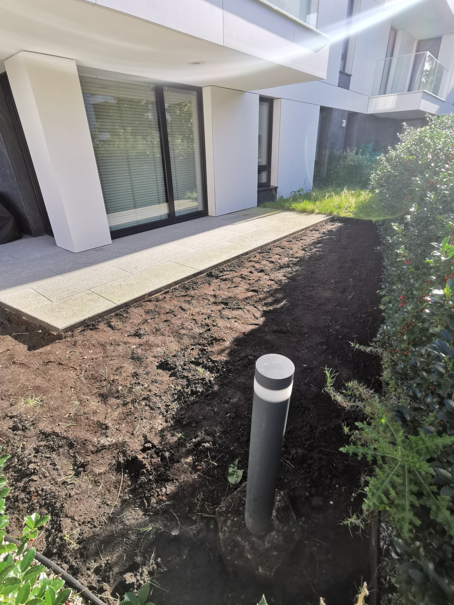 Przygotowany teren pod taras kompozytowy, widoczne obrzeża z kostki i świeżo przekopana ziemia, czarna lampa ogrodowa, tło stanowi budynek mieszkalny z dużymi oknami.