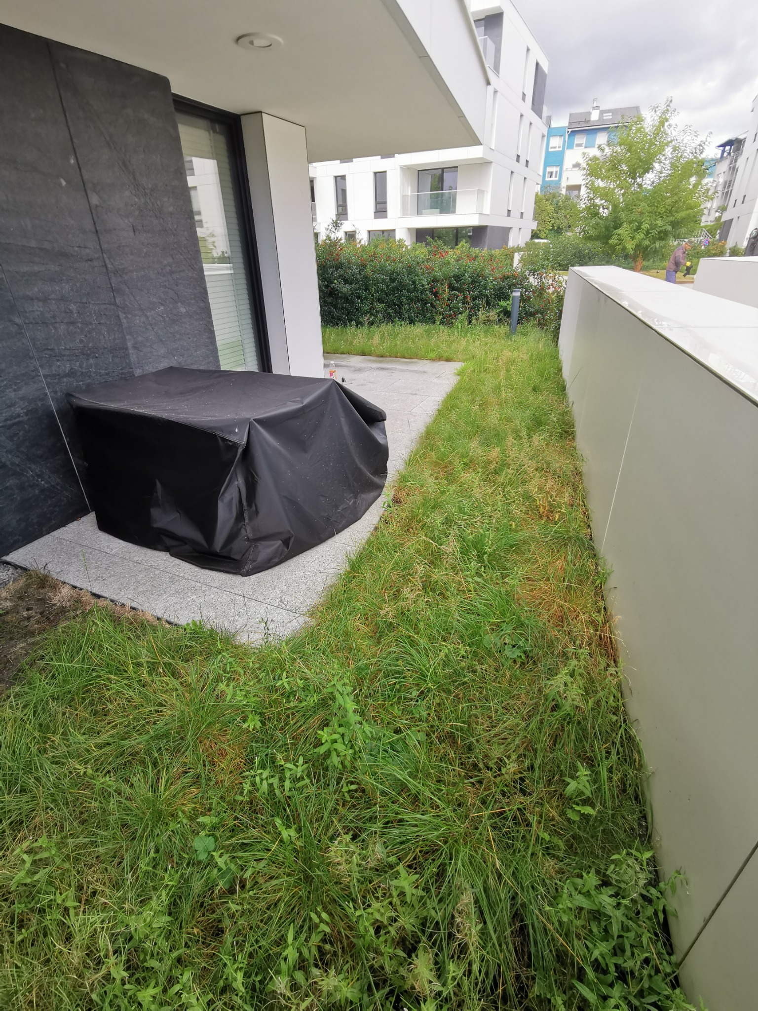 Czarne przykrycie na tarasie, obok zarośnięty trawnik i nowoczesny budynek w tle.