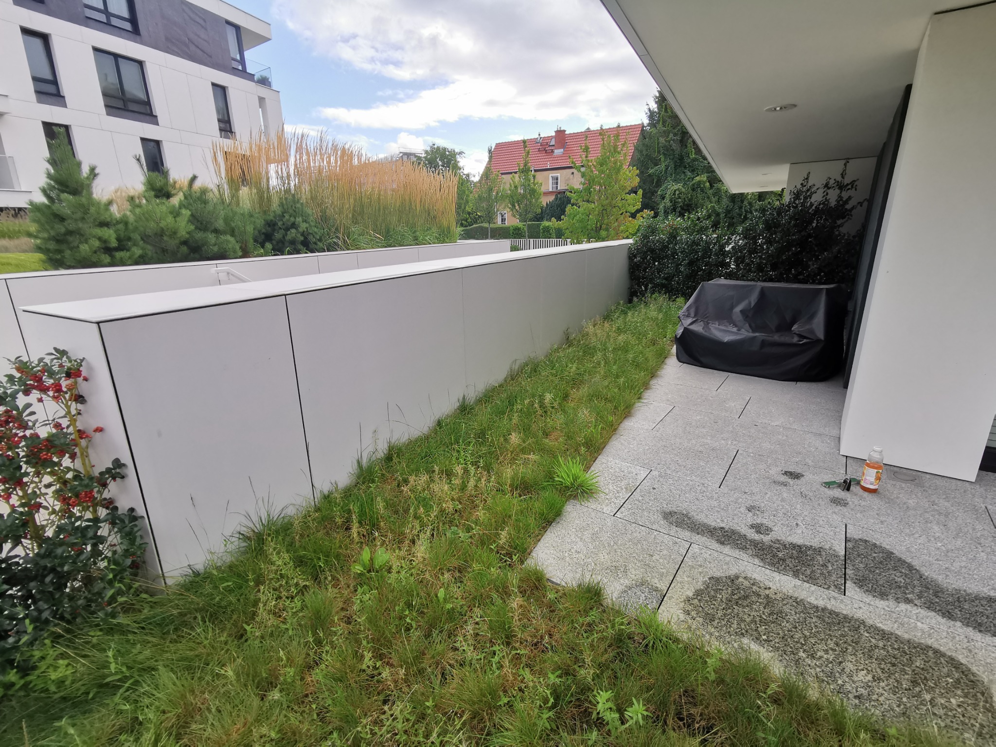 Nowoczesny taras z szarej kostki brukowej i niską białą ścianą, za którą widać trawy ozdobne i iglaki; na tarasie leżą klucze i stoi butelka z napojem.