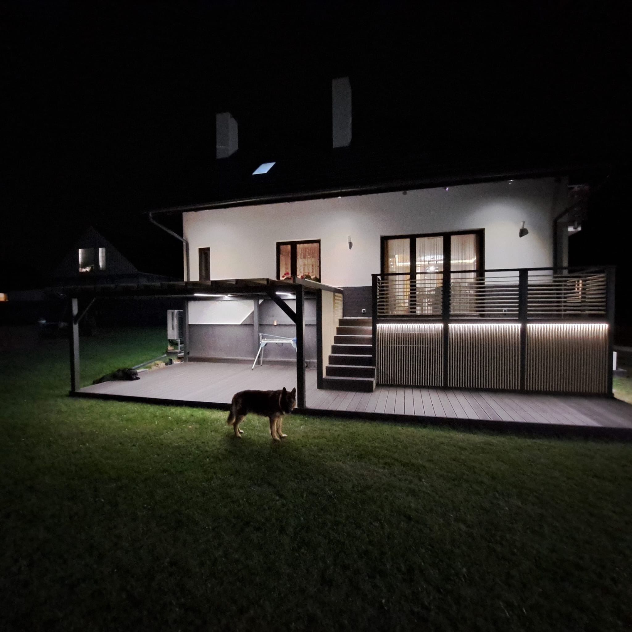 Oświetlony taras z kompozytu przy nowoczesnym domu nocą, z psem na trawniku i zadaszeniem z boku.