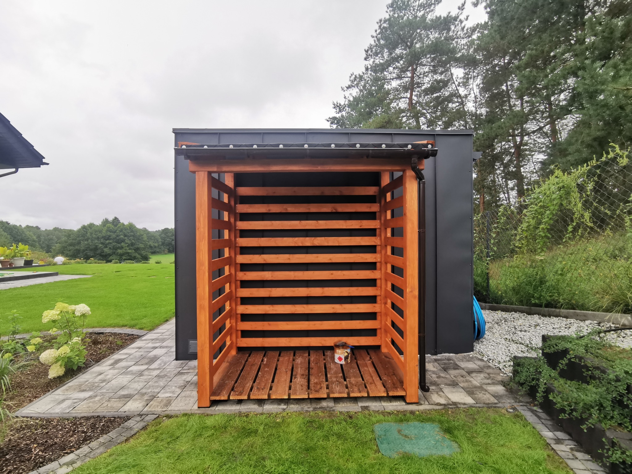 Drewniana konstrukcja, prawdopodobnie drewutnia, z pomalowanymi na brązowo elementami, stojąca na kostce brukowej, z puszką farby w środku, obok ciemnego budynku gospodarczego i zielonego trawnika.
