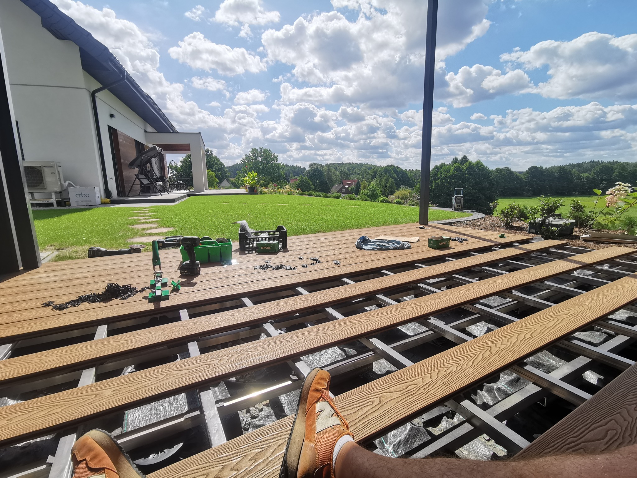 Montaż desek tarasowych na aluminiowej konstrukcji, widok z perspektywy osoby siedzącej na tarasie, narzędzia i akcesoria budowlane w tle, nowoczesny dom z zielonym trawnikiem i drzewami w tle.
