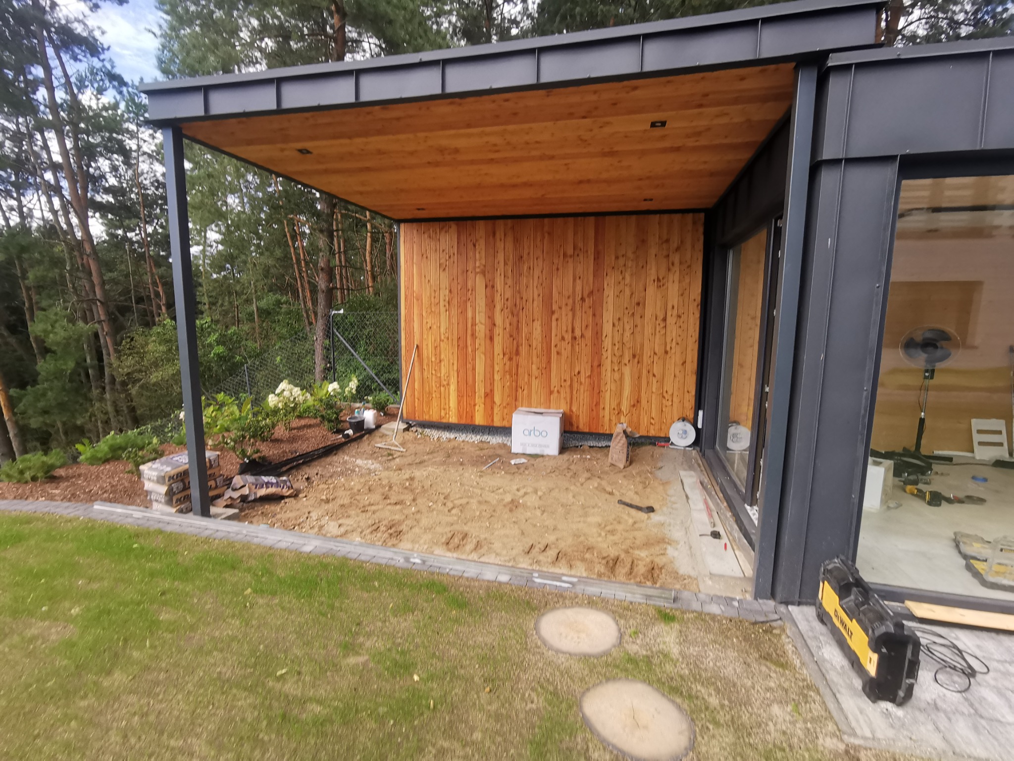 Budowa zadaszonego tarasu z drewnianą ścianą, widoczne przygotowane podłoże, narzędzia i materiały budowlane, w tle drzewa.