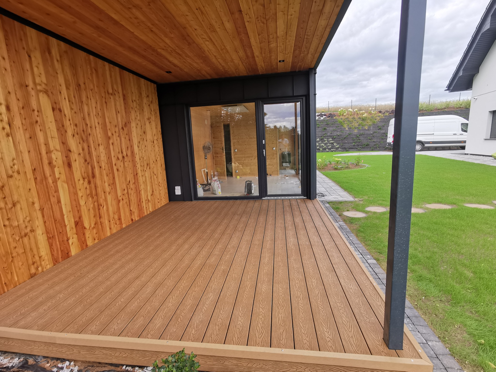 Nowoczesny taras z desek kompozytowych w kolorze jasnego drewna, przylegający do budynku z drewnianą elewacją i dużymi przesuwnymi drzwiami, otoczony zielonym trawnikiem i elementami małej...