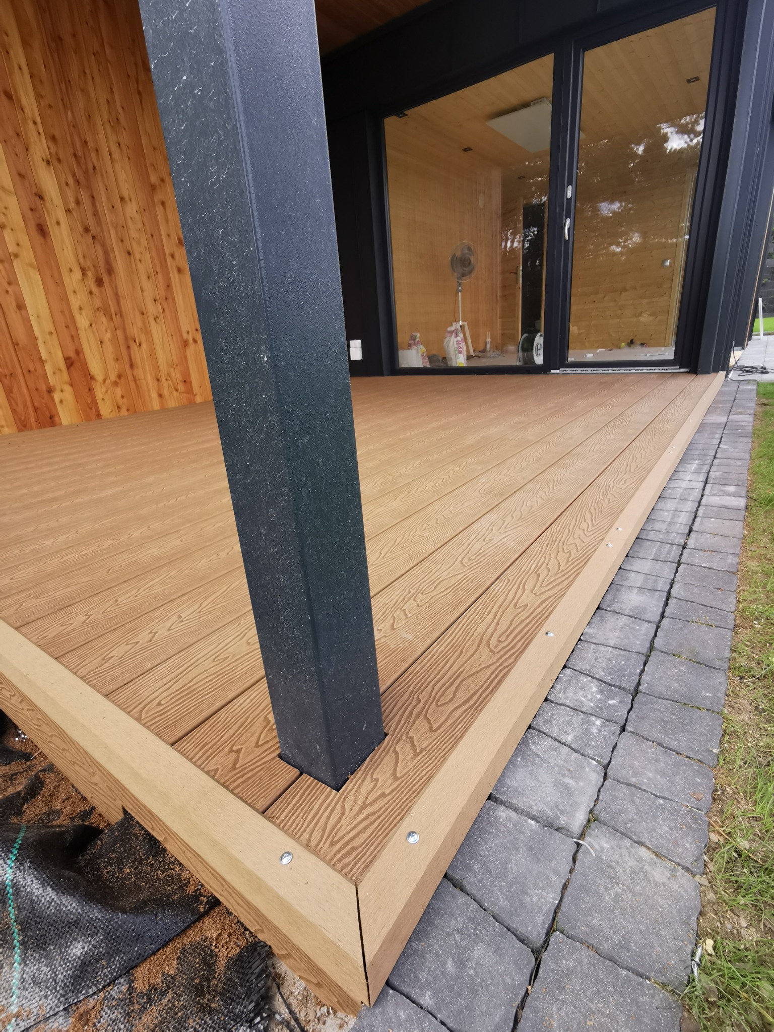 Gotowy taras z desek kompozytowych w kolorze jasnego drewna, z czarną podporą i wykończeniem kostką brukową, widok na wnętrze budynku z drewnianym wykończeniem.