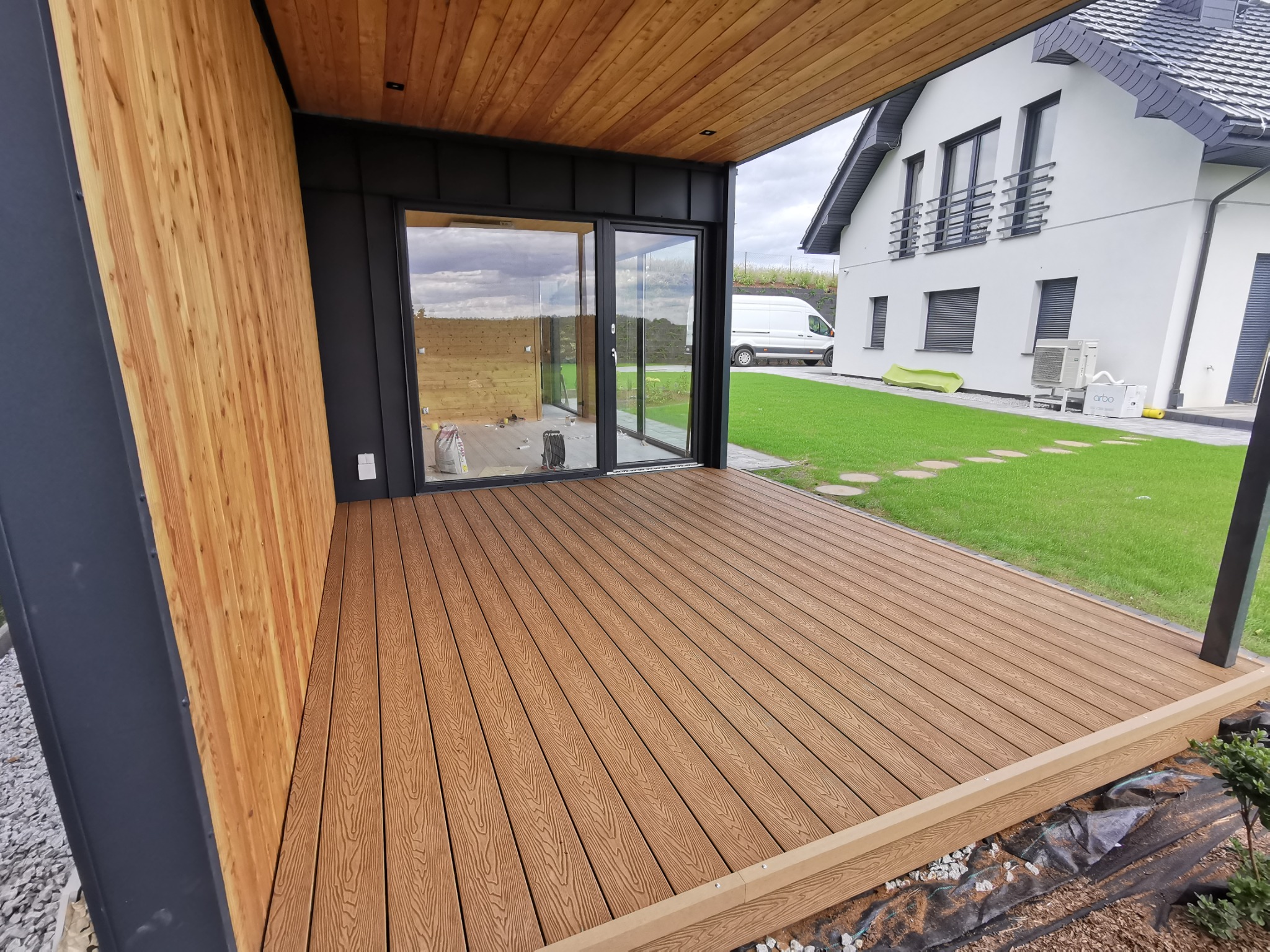 Nowoczesny taras z deski kompozytowej w odcieniach brązu, z widoczną ścianą wykończoną drewnem, czarną stolarką okienną i przesuwnymi drzwiami, na tle zadbanego trawnika i białego domu.