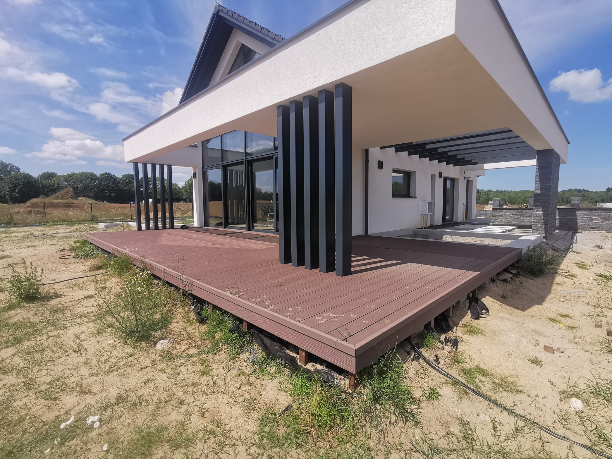 Nowoczesny dom z przestronnym tarasem z desek kompozytowych w kolorze brązu, z czarnymi filarami podtrzymującymi zadaszenie, widok na ogród i błękitne niebo.