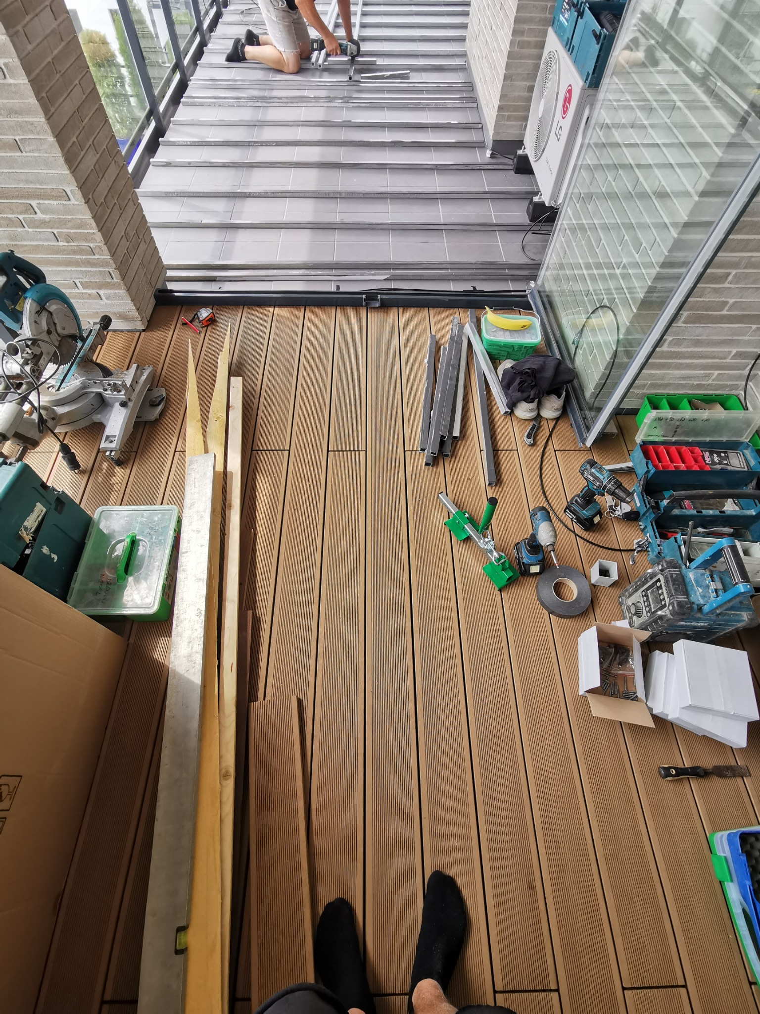 Montaż tarasu kompozytowego na balkonie: widok z góry na deski tarasowe, narzędzia, poziomice i pracownika przycinającego element konstrukcyjny.