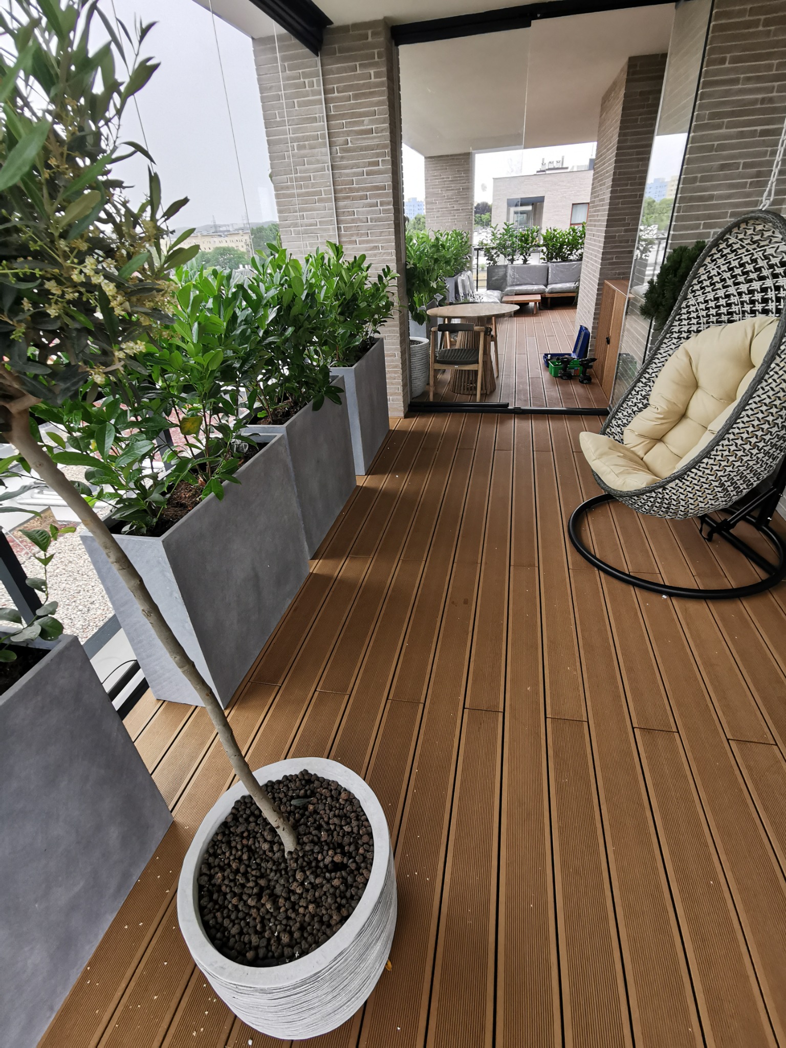 Nowoczesny taras z desek kompozytowych w kolorze ciepłego brązu, z szarymi donicami z roślinami, wiszącym fotelem typu kokon i widokiem na miasto w tle.