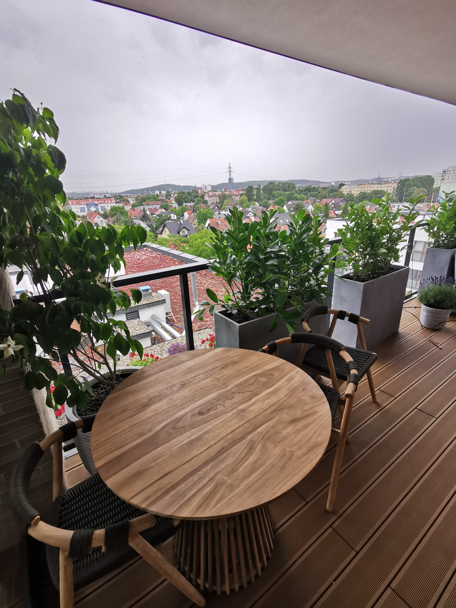 Drewniany okrągły stół i krzesła z plecionym siedziskiem na balkonie z podłogą z desek kompozytowych, w tle widok na miasto i pochmurne niebo.