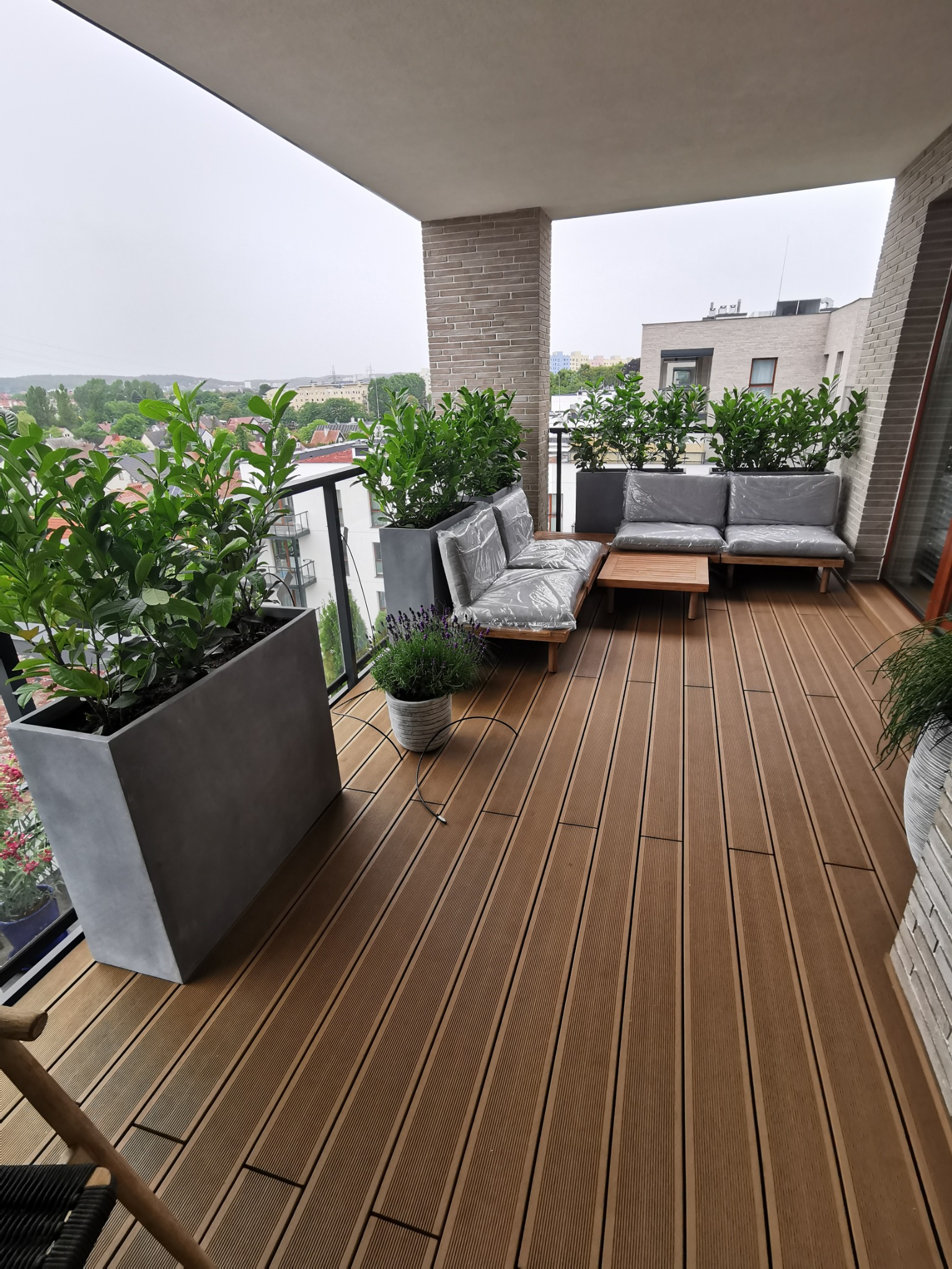 Zadaszony balkon z brązową podłogą, szarą sofą ogrodową pod pokrowcem, drewnianym stolikiem kawowym i dużymi donicami z zielonymi roślinami oraz widokiem na miasto w tle.