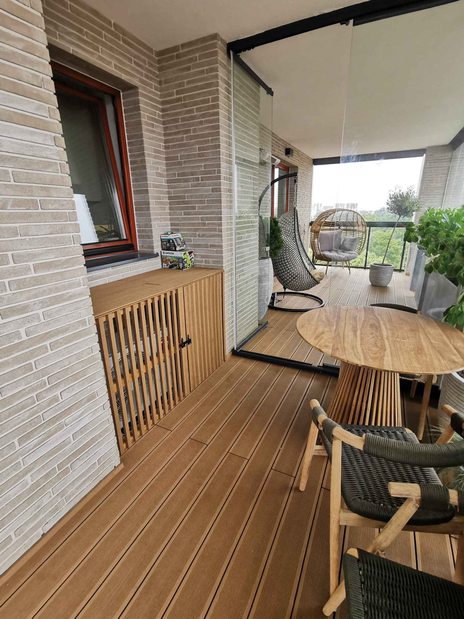 Zadaszony balkon z podłogą z desek, wyposażony w drewniany stół z krzesłami, wiszące fotele i szafkę z żaluzjowymi drzwiczkami, na której stoją dwa pudełka.