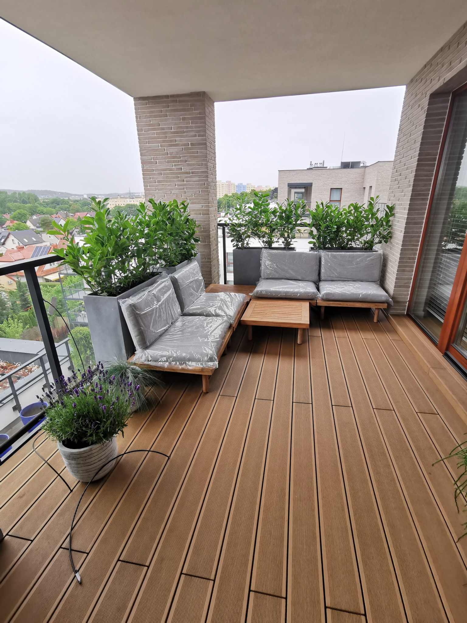 Nowoczesny balkon z brązową podłogą, meblami ogrodowymi zabezpieczonymi folią, donicami z zielenią i widokiem na miasto w tle.