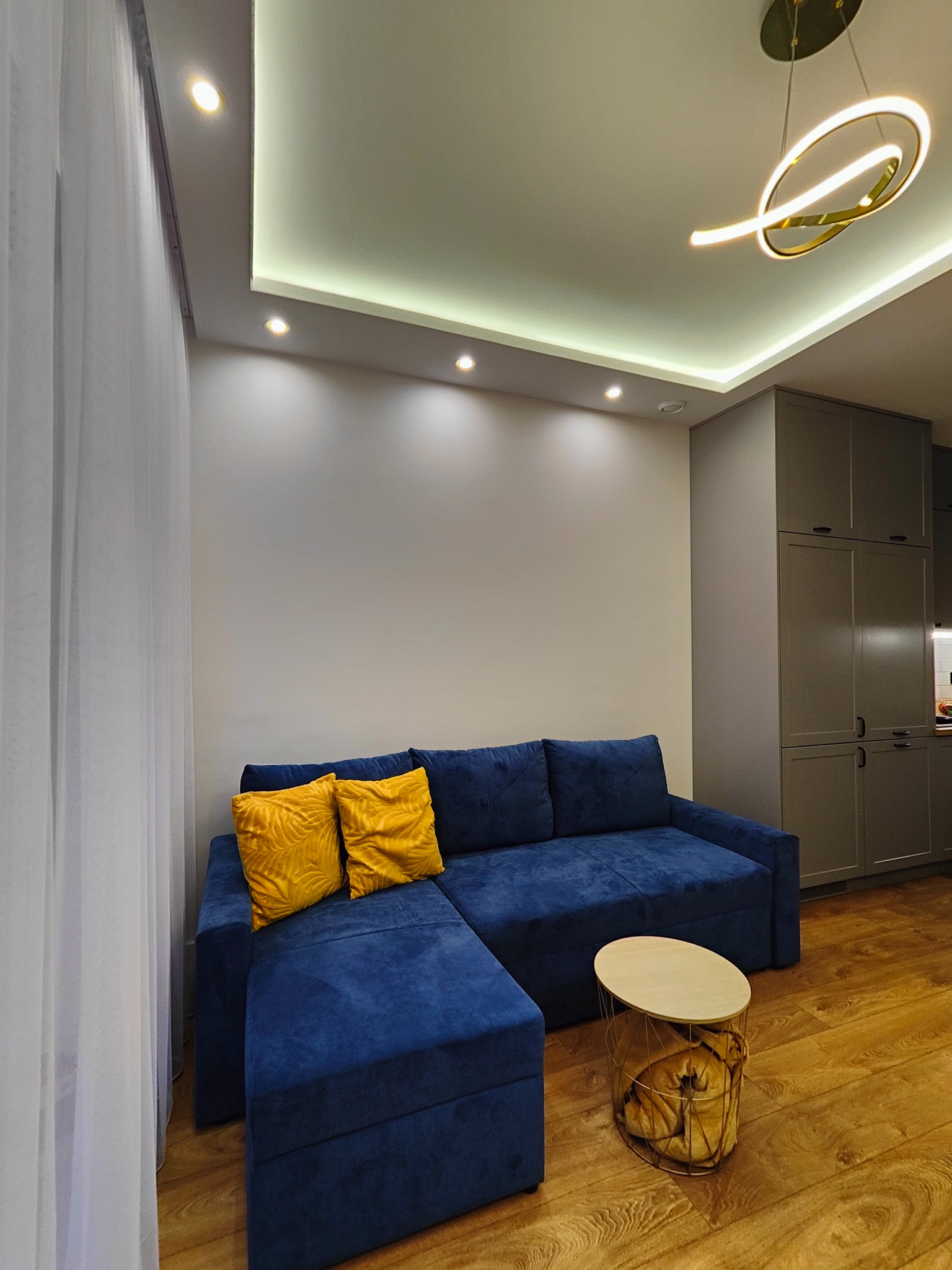 Nowoczesny salon z narożną sofą w odcieniach granatu, żółtymi poduszkami, okrągłym stolikiem kawowym z metalową podstawą i drewnianym blatem, parkietem oraz oświetleniem sufitowym LED.