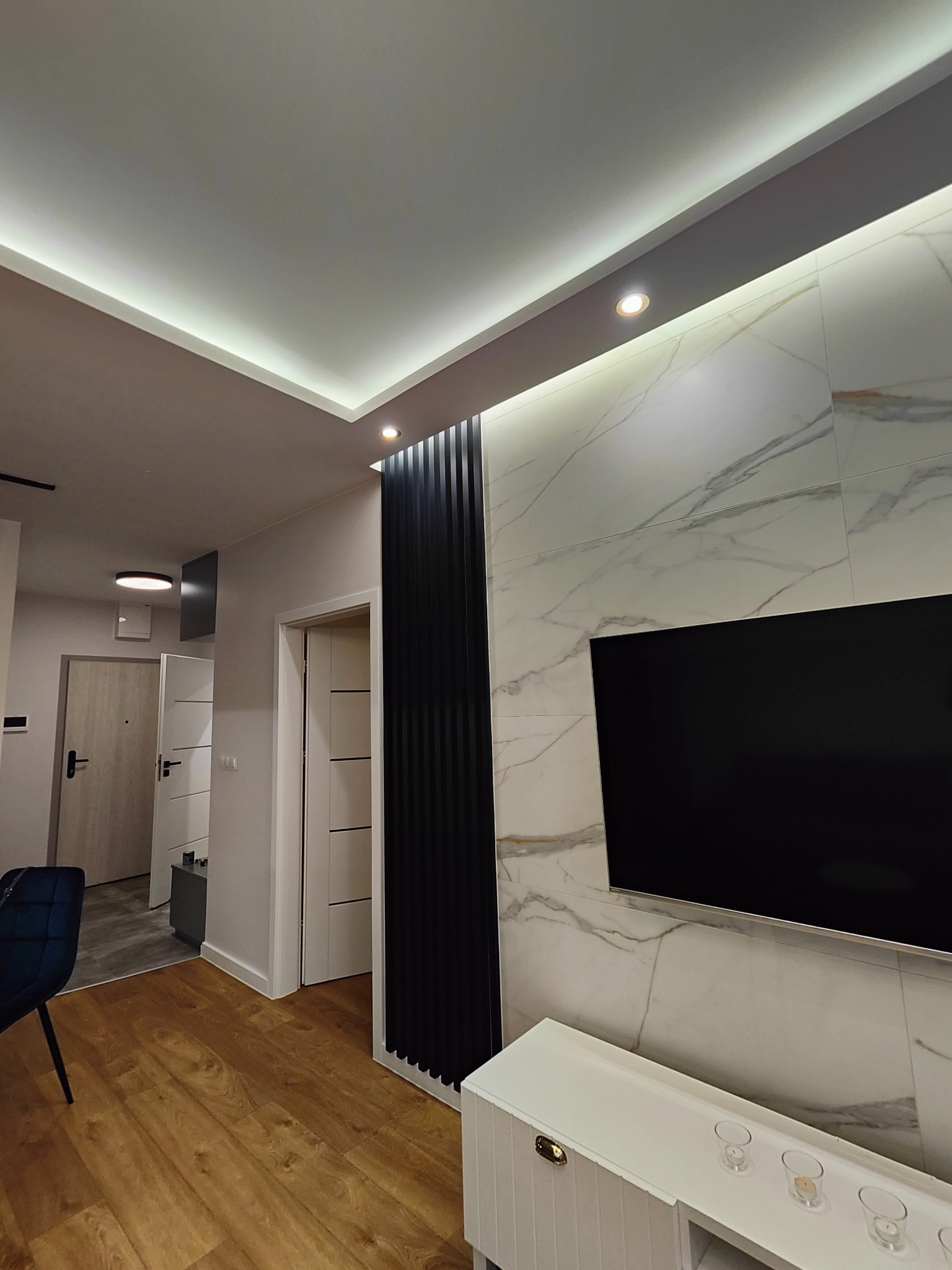 Nowoczesne wykończenie wnętrza salonu z marmurową ścianą, panelem dekoracyjnym i podświetlanym sufitem, widok na korytarz z drzwiami.
