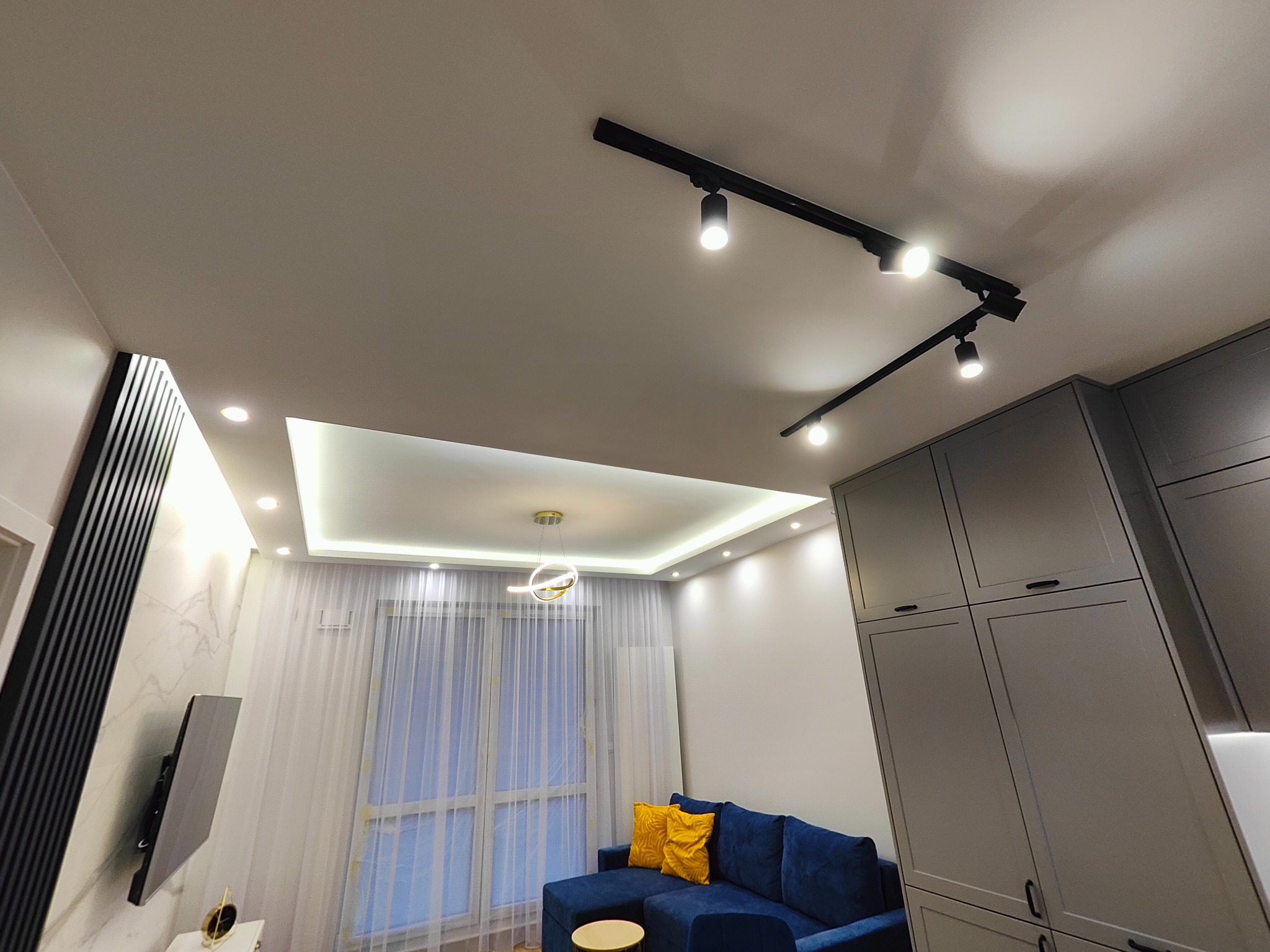 Nowoczesny salon z niebieską sofą, żółtymi poduszkami, szarą zabudową meblową, oświetleniem szynowym i podwieszanym sufitem z oświetleniem LED.