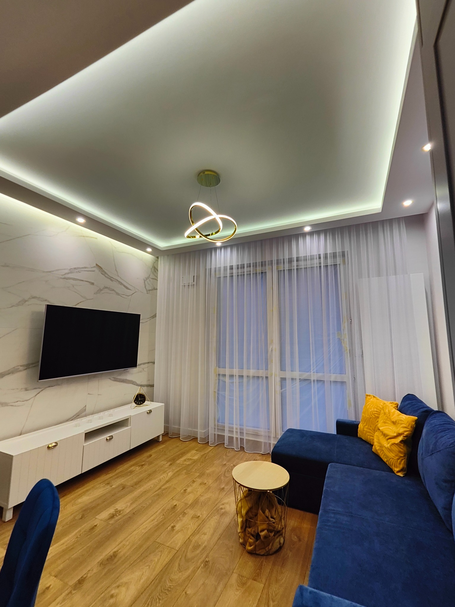 Nowoczesny salon z niebieską sofą narożną, marmurową ścianą z telewizorem, białym stolikiem RTV, okrągłym stolikiem kawowym ze złotymi akcentami i designerską lampą sufitową.