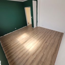 Adrian Stando - Pokój po remoncie z ułożonymi panelami podłogowymi w odcieniach brązu, ściana pomalowana na ciemnozielony kolor, widoczne niedociągnięcia malarskie przy podłodze.