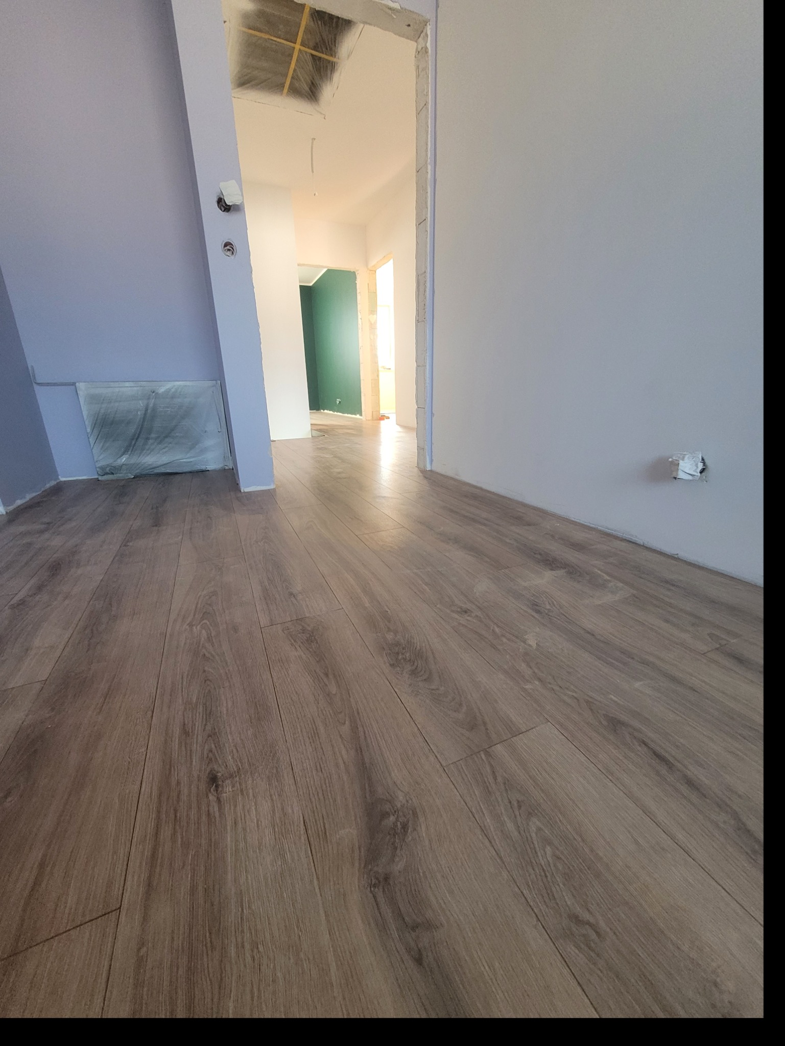 Ułożone panele podłogowe w trakcie remontu, widoczne ściany w kolorze fioletowym i szarym, zabezpieczona folią grzejnik, surowy otwór drzwiowy i fragment zielonej ściany w głębi.